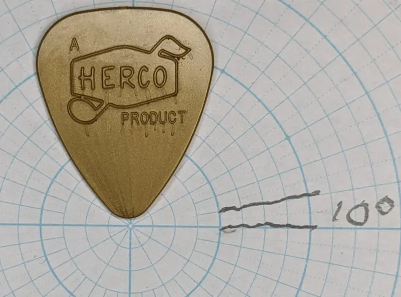 The Gold Herco Vintage Reissue pick against circular angle ruled paper for shape reference. Note the textural relief embossing, seen on both sides.