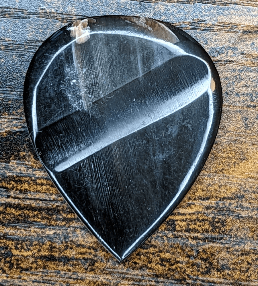 The GuitarHeads' Bufflo Horn pick in black against a woodend backdrop revealing the grain of the pick material. Note the grip indent.