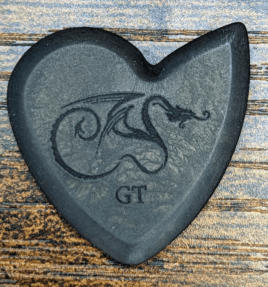 The Dragon's Heart GT Pick Viewed Against Wood Grain Background For your viewing pleasure. Note the dark black color and the Engraved Dragon.