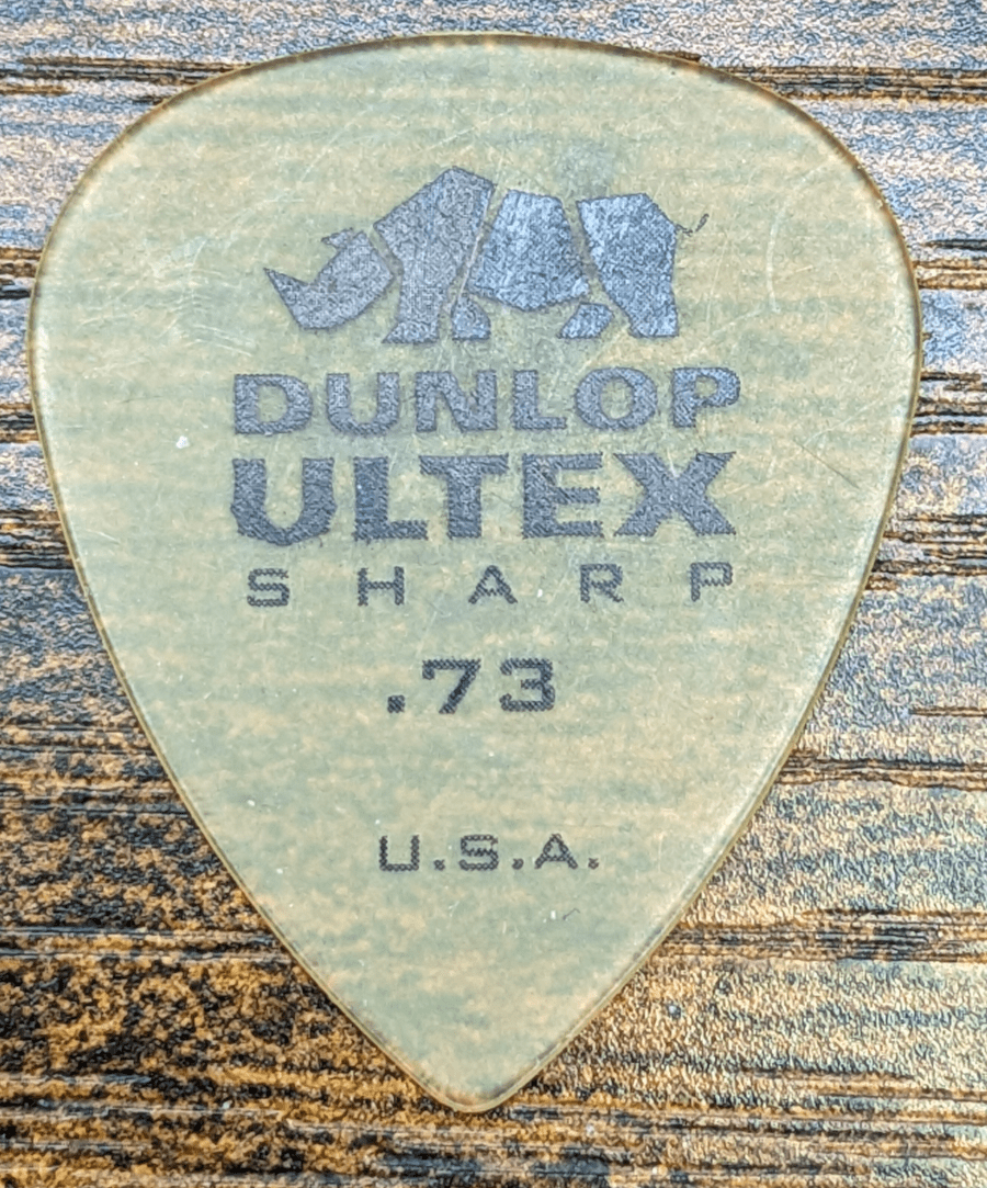 Jim Dunlop's Ultex Sharp pick 0.73mm set on wood background. Note the rounded fine tip for speed playing.