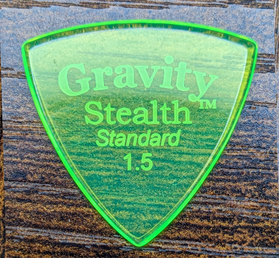 The Gravity Brand Stealth Standard 1.5 Green Pick: A glowing Green Demon, seen against a brown and tan wood backdrop. Note Fluourecsence!